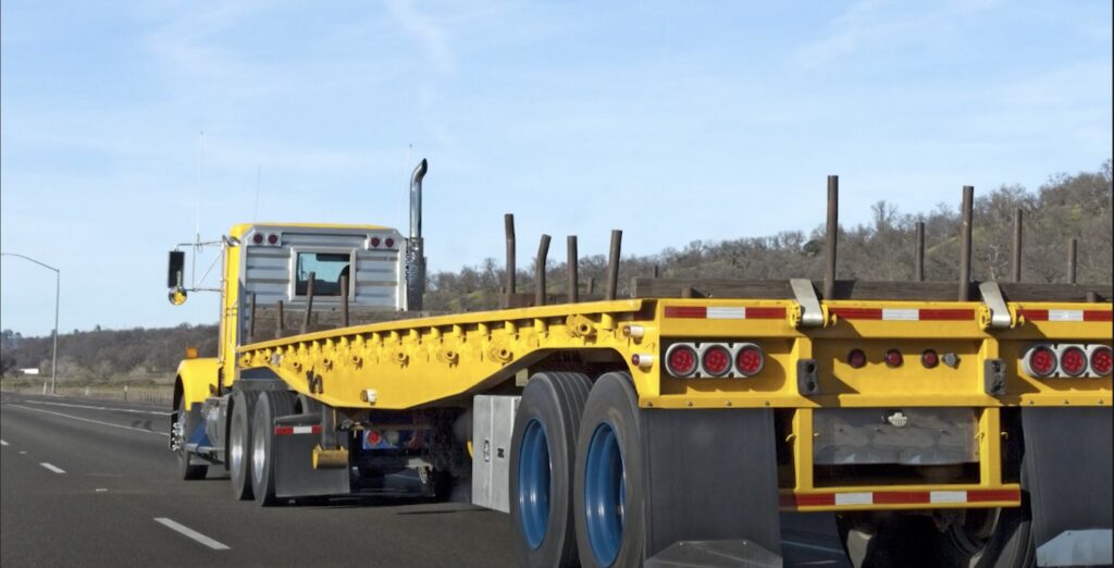 Image shows an empty, yellow flatbed trailer driving on the highway.