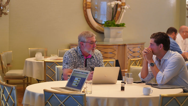 Two men are sitting at a round, white table with laptops having a discussion within a delightfully decorated restaurant.