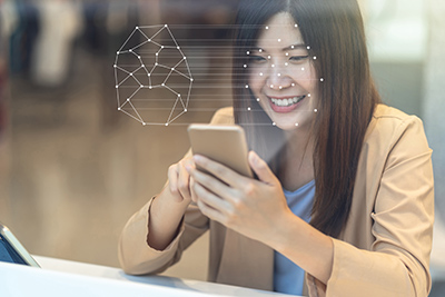 An Asian woman with long brown hair sits at a desk, smiling, using a smartphone and its facial recognition application.