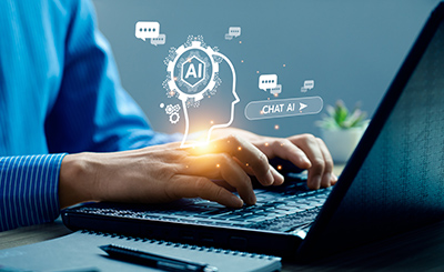 A white person in a blue shirt sits at a desk with a laptop open, hands resting on the keyboard like they are about to type. Graphics are above the hands that read "Chat AI", representing the person interacting with Generative AI.