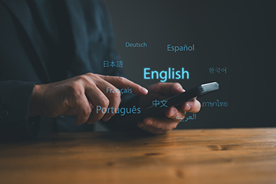 A white person is sitting at a wooden desk, holding a smartphone with one hand, and using the other hand to use it. Surrounding the person's hands and phone are several different languages represented, including English, Espanol, Deutsch, and more - showcasing a translation application as an example of Natural Language Processing.