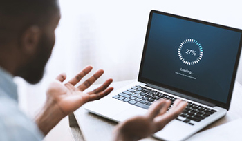 A black man is sitting at a desk with a laptop on it. The laptop is open and shows a blue loading screen while the man has his hands up in frustration. 