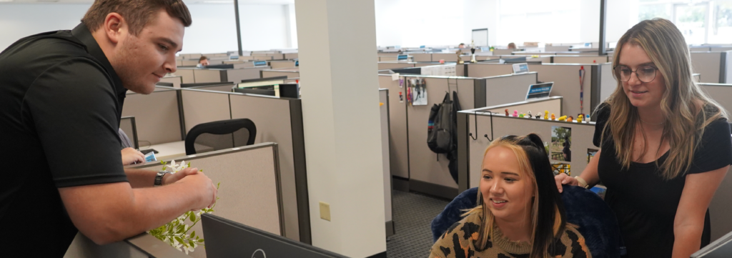 A photo of Trinity Team Members. A young, white woman with long dark hair is sitting at a desk staring at a computer screen. Standing behind her is another young, white woman with long blond hair and glasses wearing a black t-shirt, smiling and also looking at the computer screen. Both girls are inside a cubicle with a background of being in an office full of cubicles. Standing in front to the left of the women, leaning on the cubicle wall and overlooking onto the computer screen is a young, white male with short dark hair and wearing a black polo shirt. 