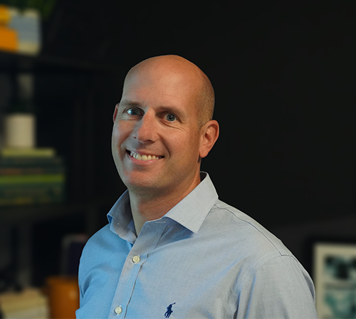 Pictured is a headshot of Chris McAvoy, Director of Managed Services at Trinity Logistics. Chris is a white adult male, bald, blue eyes, smiling, and wearing a light blue button down shirt in front of a dark background. 