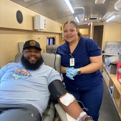 Photo of young, black man, with a beard, wearing a baseball cap and light blue graphic t-shirt, sitting inside of a mobile blood drive, giving blood, and smiling. Standing besides him, also smiling and with her hands clasped at her waist, is a young tan-skinned female nurse, with long dark hair that is tied back, wearing dark blue scrubs, her badge, blue latex gloves, and a smartwatch on her left wrist.