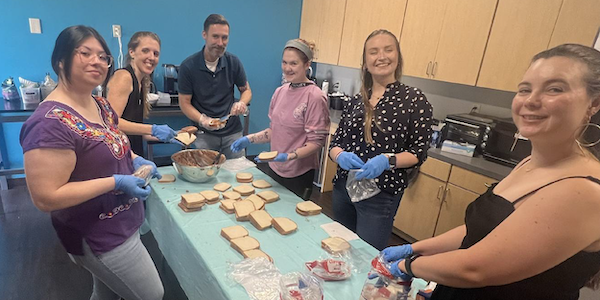 A photo showing Texas Trinity Team Members preparing peanut butter and jelly sandwiches for the homeless. 6 Team Members are pictured around a table with a light blue tablecloth on it, along with a bowl of the pb&j mix, slices of bread, and sandwiches in bags. All are looking at the camera and smiling. From left to right: a middle-aged tan skinned woman with long black hair tied back, rose-rimmed glasses, wearing a short-sleeve purple blouse with colorful accents around the front neckline, wearing blue gloves, holding a packaged sandwich; a middle-aged white woman, with long strawberry blond hair tied back, wearing a sleeveless black blouse, blue gloves, and applying the pb&j mixture to a piece of bread; a middle-aged white man, taller than the women, short brown hair and a short beard, wearing a navy short-sleeve buttoned polo, clear gloves, also slathering on pb&j mixture to bread; a shorter young white woman, long dark blond hair tied up with a headband, wearing a long sleeve light pink shirt with the sleeves rolled up to her elbows, blue gloves, and holding a piece of bread; a taller young white woman with long blond hair, half up, her eyes are closed as she's smiling, wearing a black long sleeve blouse with white polka dots, blue gloves, holding an empty bag; and a young white woman with long brown hair tied back, hoop earrings, wearing a black spaghetti strap top, blue gloves, and holding a packaged loaf of bread. Behind them are light brown colored cabinets on the right and a blue wall on the left.