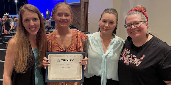 A photo of Trinity Team Members standing and smiling for a picture with one of the scholarship winners. From left to right: a middle-aged white woman with long strawberry blond hair, wearing a sleeveless blue tank top with a sleeveless black vest over top of that; a young high school graduate, white girl, wearing clear rimmed glasses, long blond hair that is tied back, wearing a short sleeve flowy and flowery pattern dress, holding a certificate with the Trinity logo on it; a young white woman with long dark brown hair tied up and wearing a light blue long sleeve blouse with a tie in the middle; a middle aged white woman with long blond and gray hair tied back with a red headband, black rimmed glasses, long colorful earrings, and a black short sleeve t-shirt that reads 'blessed'. Behind them looks to be a background of an auditorium.
