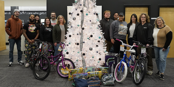 A photo of Trinity Team Members standing and smiling at a camera, group up on either side of a white decorated Christmas tree with unwrapped presents like bikes and legos in front of the tree. There are six people on each side of the tree, smiling, and a mix of young and middle aged, women and men, white and black.