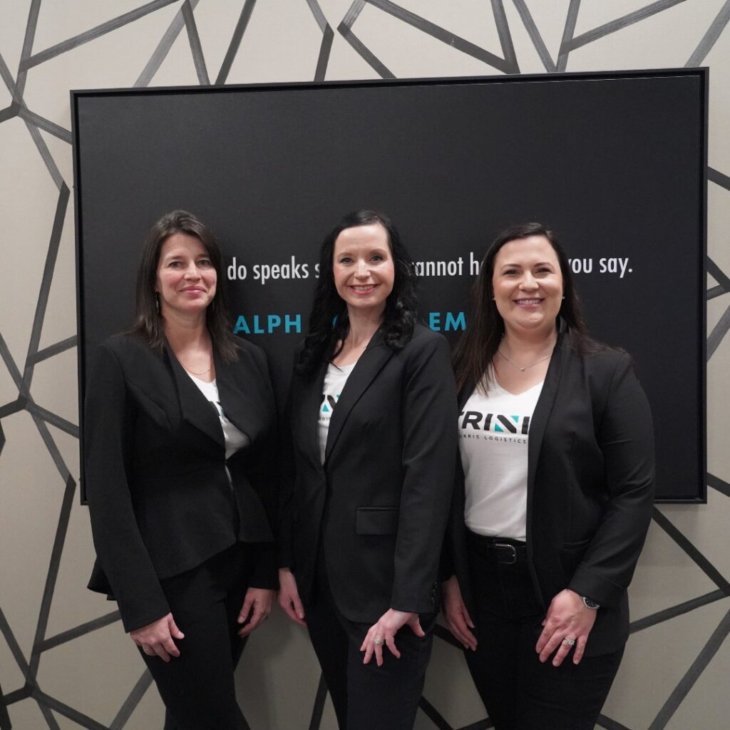 Pictured left to right are the three members of the Fraud Force team, Tracy Mitchell, a middle aged white woman with long dark brown hair, wearing a white t-shirt with the Trinity logo and a black blazer over top, dark pants, smiling; then Kristin Deno, a middle aged white woman with long black hair, wearing the same, then Angela Smith, a young white woman with long dark brown hair, wearing the same. The group stands in front of one of Trinity's patterned walls, tan with black abstract lines all over, and a framed quote on black canvas and white text.