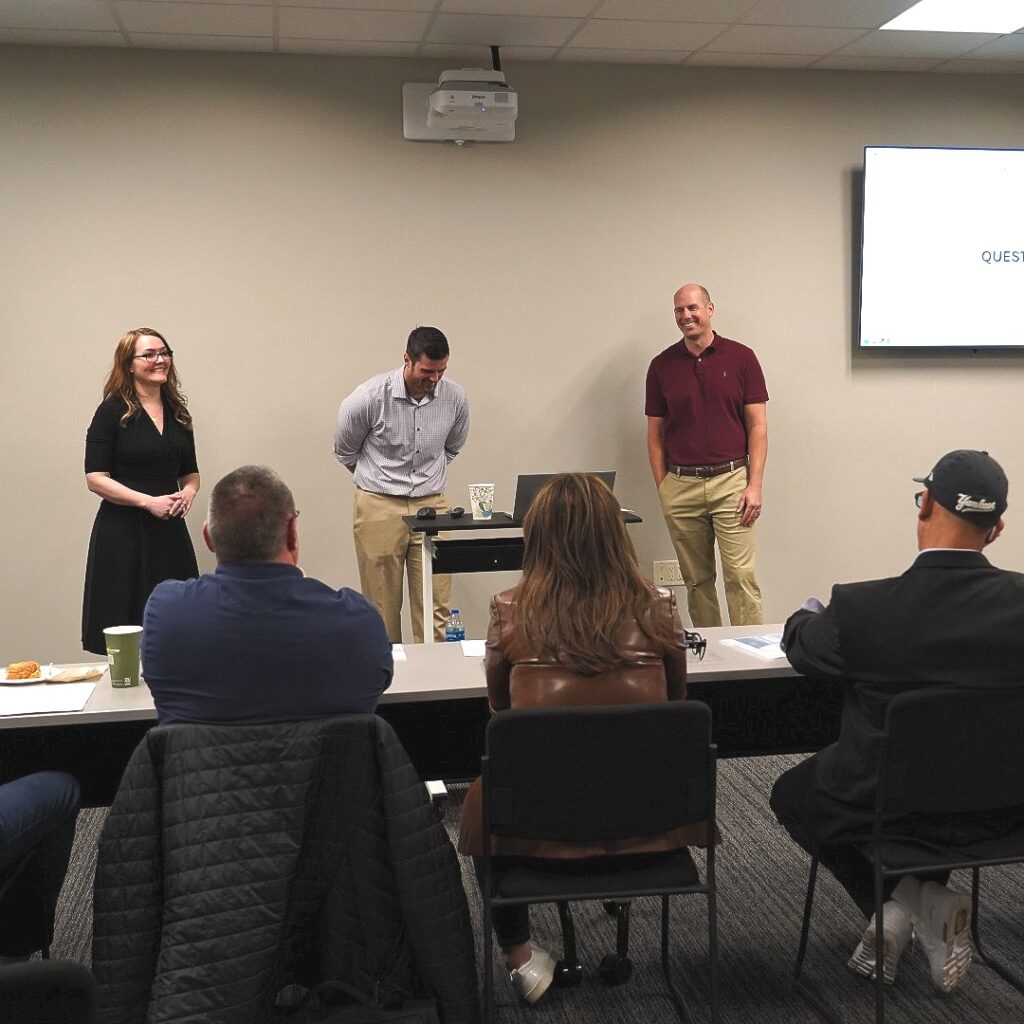 A photo the Predictive Profits group, taken during their pitch. From left to right is Cheryl Swain, a young white woman with long blond hair, black rimmed glasses, smiling, wearing a black long sleeve dress. To the right of her is Zach Howard, standing in front of a podium with his laptop. He is a young white man, with short dark hair, smiling, perhaps laughing, slightly bent over, clean shaven, wearing a light colored button up shirt, long sleeve, and khakis. To the right of him and the podium is Chris McAvoy, smiling or laughing as well, a middle aged white man, bald and clean shaven, wearing a maroon red short sleeve polo shirt and khakis with a dark brown belt. To the right of him is a small glimpse of a large screen, with their presentation, mostly white with some black text. In front of the group you can see the back of heads of some of the judges sitting in front of a table with paper and pens in front of them.