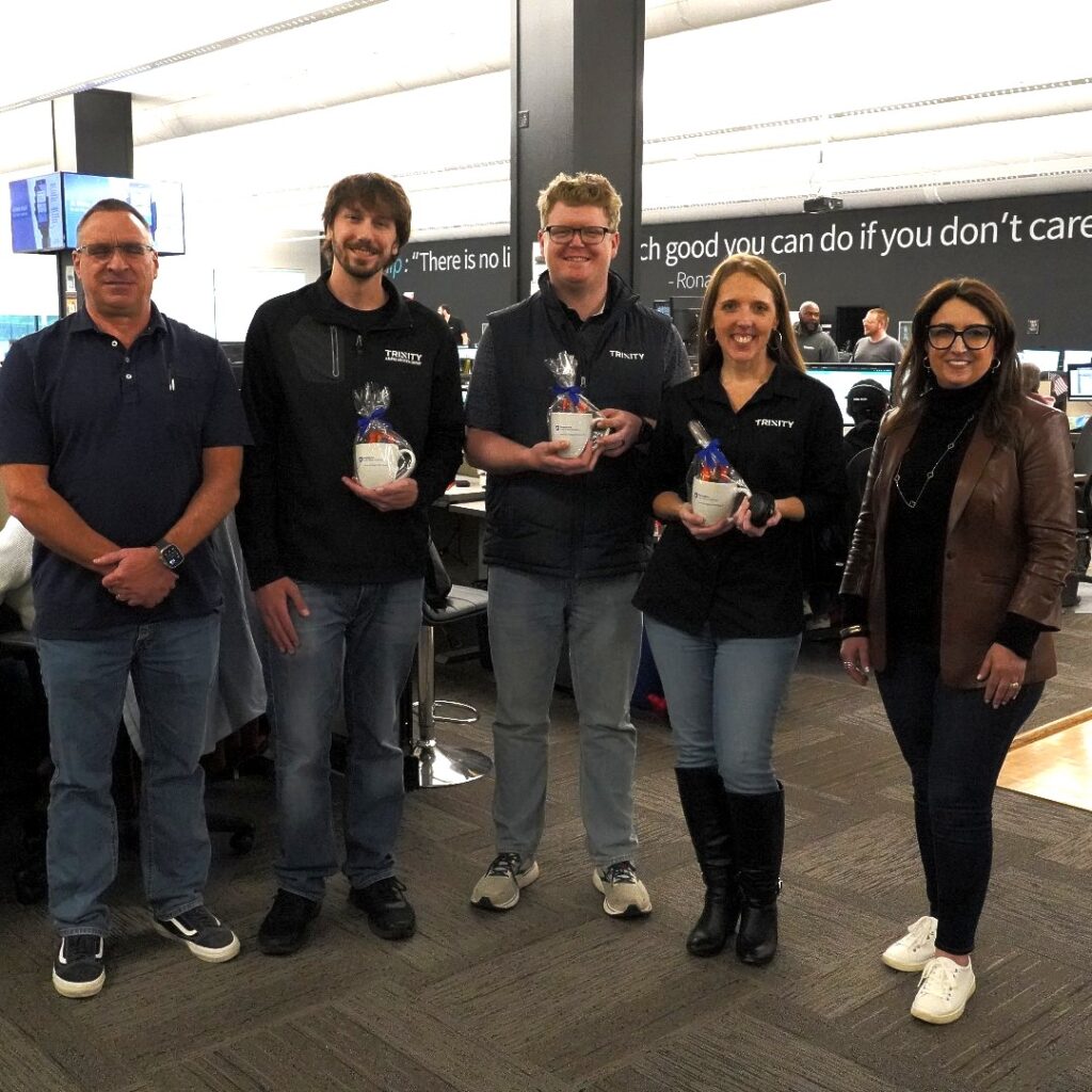 A picture of the winners of the Week of Wonder competition, the Trinity CarrierOne Experience, along with two of the judges. From left to right stands Donnie Burris, a middle-aged white man with short dark hair, glasses, clean shaven and smiling, wearing a navy polo t-shirt and blue jeans with sneakers; James Beveridge, Trinity Team Member and part of the CarrierOne group, a young white man with shaggy dark brown hair, a short beard and mustache, smiling, wearing a black long sleeve zip up with the Trinity logo, blue jeans, and black sneakers, holding a gift set with a white mug and other goodies wrapped in cellophane; Michael Adams, a Trinity Team Member and part of the CarrierOne group, a young white man with short blond hair, glasses, clean shaven and smiling, wearing a dark t-shirt with a black zip up vest with the Trinity logo over that and blue jeans with white sneakers, holding his prize, the same as James; Amanda Ashford, Trinity Team Member and part of the CarrierOne group, a young white woman with long red hair, smiling, wearing white hoop earrings, a long sleeve black zip up with the Trinity logo, blue jeans and knee high black boots, holding the same prize as the others; and Donna Palumbo-Miele, the Executive Director for the Center for Supply Chain Research at Penn State, a young white woman with long dark brown hair, smiling, wearing black rimmed glasses, hoop earrings, a black long sleeve turtleneck with a brown leather blazer over top, a long necklace, dark jeans, and white sneakers, along with several rings. Behind them is the Trinity Operations floor consisting of Team Members working at their desks.