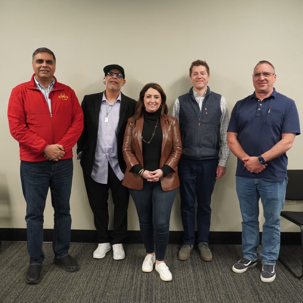 This photo shows the judges, standing and smiling for the camera. Left to right is Dr. Raj Agnihotri, a medium brown skinned middle aged man, with short dark gray hair, clean shaven, wearing a long sleeve red zip up jacket with the Iowa State logo on the right breast and a light colored button up shirt with a collar underneath, dark blue jeans, and black loafers, hands crossed in front of him; Joe Negron a middle aged light brown skinned man, wearing a black baseball cap, sunglasses, a bluetooth ear piece on his right ear, a light blue gray button up dress shirt, untucked, a black blazer over top, possible dog tags, dark slacks, and white sneakers, leaning back with his hands in his pockets; Donna Palumbo-Miele, a middle aged white woman with long brown hair, wearing hoop earrings, a black long sleeve turtleneck with a brown leather blazer overtop, a long necklace, gold bangles on her right wrist, dark blue jeans, white sneaks, hands crossed in front of her; Zach Buckner, a middle aged white man with short brown hair, clean shaven, wearing a plaid striped but light button up dress shirt, a gray zip up vest over top, dark blue jeans, dark gray sneakers or loafers, with his hands by his sides, and Donnie Burris, a middle aged white man, wearing glasses, clean shaven, short cropped dark hair, a navy blue polo t-shirt, blue jeans, and dark sneakers, with his hands crossed in front of him.
