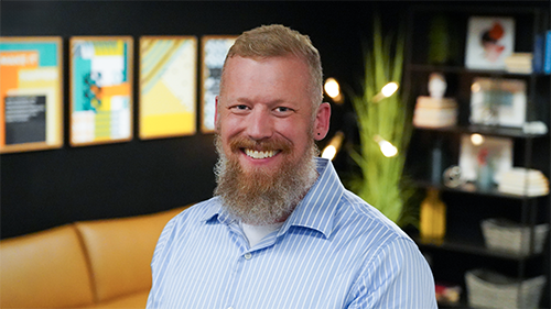 Photo of Jason Varland, Director of Engineering at Trinity, a smiling light-skinned toned man with short blond hair and a blond and gray beard, wearing a blue and white striped collared button down shirt. 