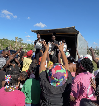 A group of people gathered around an open truck during a Hurricane Melissa relief effort in Jamaica. Several individuals on the truck are handing out bags of supplies while community members reach up to receive them under a clear blue sky.