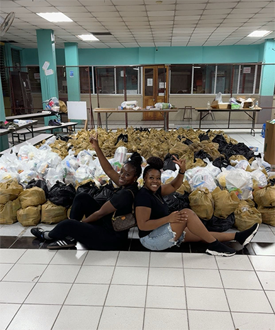 Jordan Walker and Nastacia Crooks Brady sit on the floor in front of rows of organized Hurricane Melissa relief bags in an indoor facility with teal walls and tiled flooring. Behind them, tables hold additional supplies for distribution.