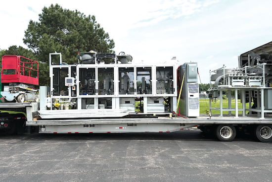 White industrial machine with multiple enclosed compartments and control panels, loaded on a flatbed trailer for freight transport, with trees and a building visible in the background.