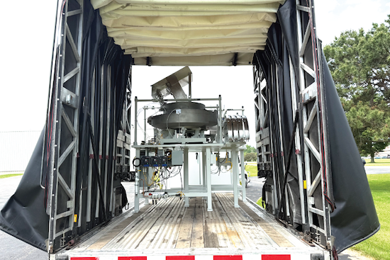 View into the open rear doors of a curtain-side trailer, showing a large stainless steel funnel-shaped piece of equipment secured on a raised platform inside.