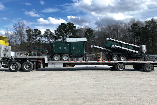 Two pieces of dark green industrial equipment, including a folded conveyor or lift arm, loaded on a flatbed trailer for transport in a gravel lot.