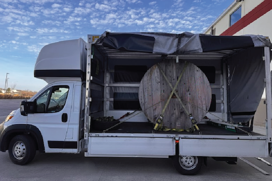 A large wooden cable spool secured with straps and chains inside the open cargo area of a box truck."