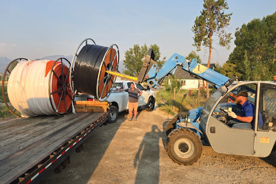 A man stands beside a flatbed trailer loaded with large cable spools while a telehandler forklift maneuvers nearby at an outdoor job site, with mountains in the background.