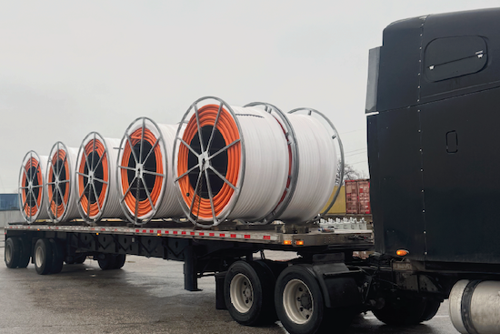 Several large spools of orange and white coiled cable or conduit loaded end-to-end on a flatbed trailer, parked behind a semi-truck cab."
