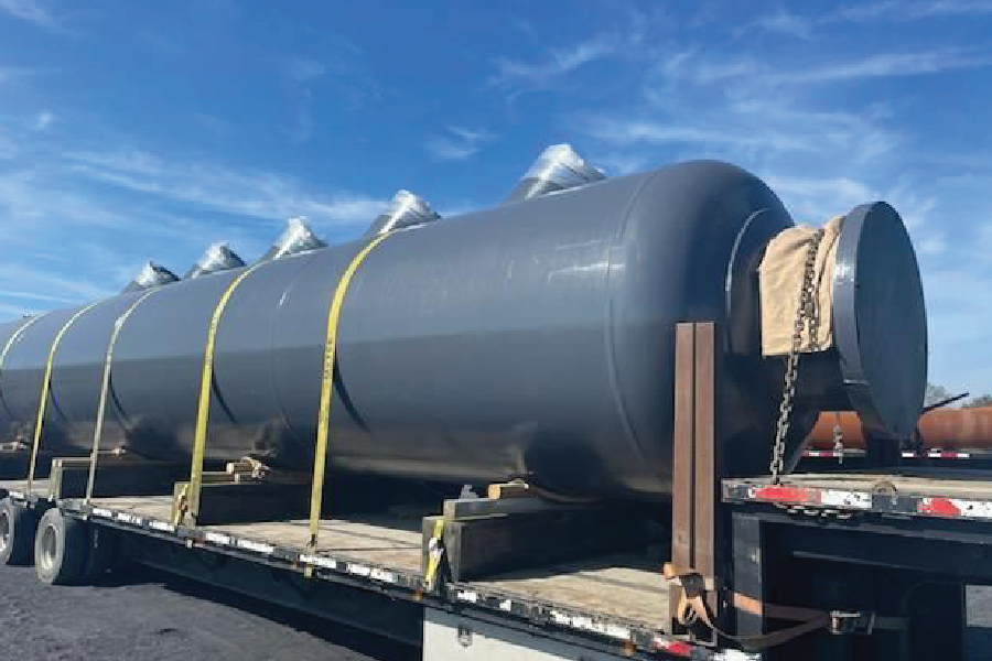 Large gray cylindrical steel tanks strapped to a flatbed trailer, loaded end-to-end for transport under a clear blue sky.