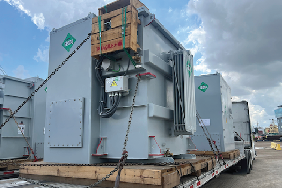 A large gray industrial transformer, secured with tie-down chains and a wooden crate marked 'Fragile' on top, loaded on a flatbed trailer for freight transport. Bright green diamond-shaped labels mark the unit's corners, and the load is chained and blocked with wood cribbing to prevent shifting during transit.
