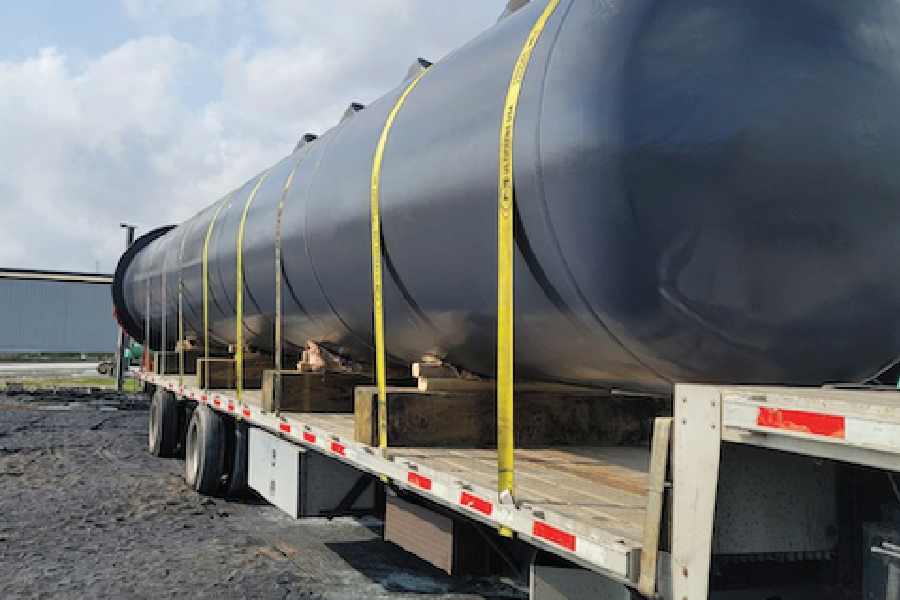 A large black cylindrical tank secured with yellow tie-down straps on a flatbed trailer, loaded on wooden blocking for freight transport. The tank extends the length of the trailer, with a paved lot and warehouse building visible in the background under a partly cloudy sky.