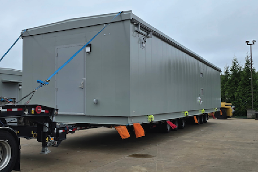 A gray modular building unit, resembling a portable office or storage structure, loaded on a multi-axle trailer for freight transport, secured with a blue ratchet strap and orange stabilizing blocks. The trailer sits in a paved lot with trees and equipment visible in the background under an overcast sky.