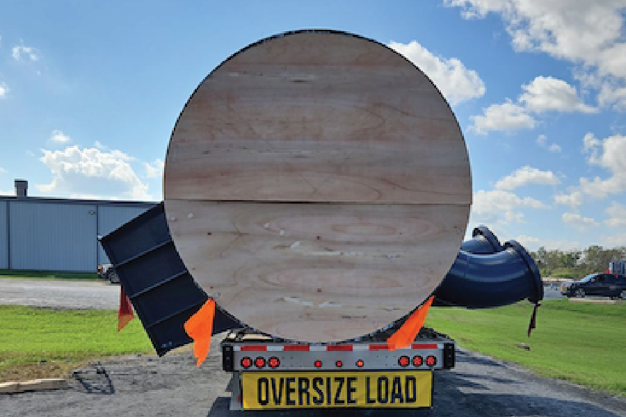 Rear view of a large cylindrical piece of equipment with a round plywood end cap, loaded on a trailer marked with an 'Oversize Load' banner and warning flags for wide-load freight transport. A dark curved component extends from the side of the load, with a warehouse building and parking lot visible in the background under a partly cloudy sky.