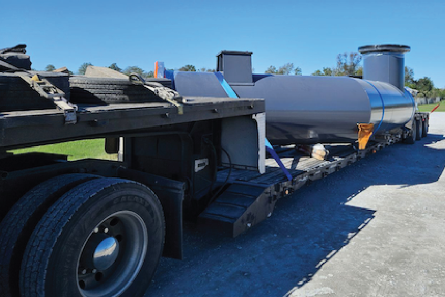 A large gray industrial tank with a cylindrical vertical stack, secured with blue straps on a flatbed trailer for freight transport. The equipment sits behind the truck's cab on a paved lot, with trees and open sky in the background on a clear day.